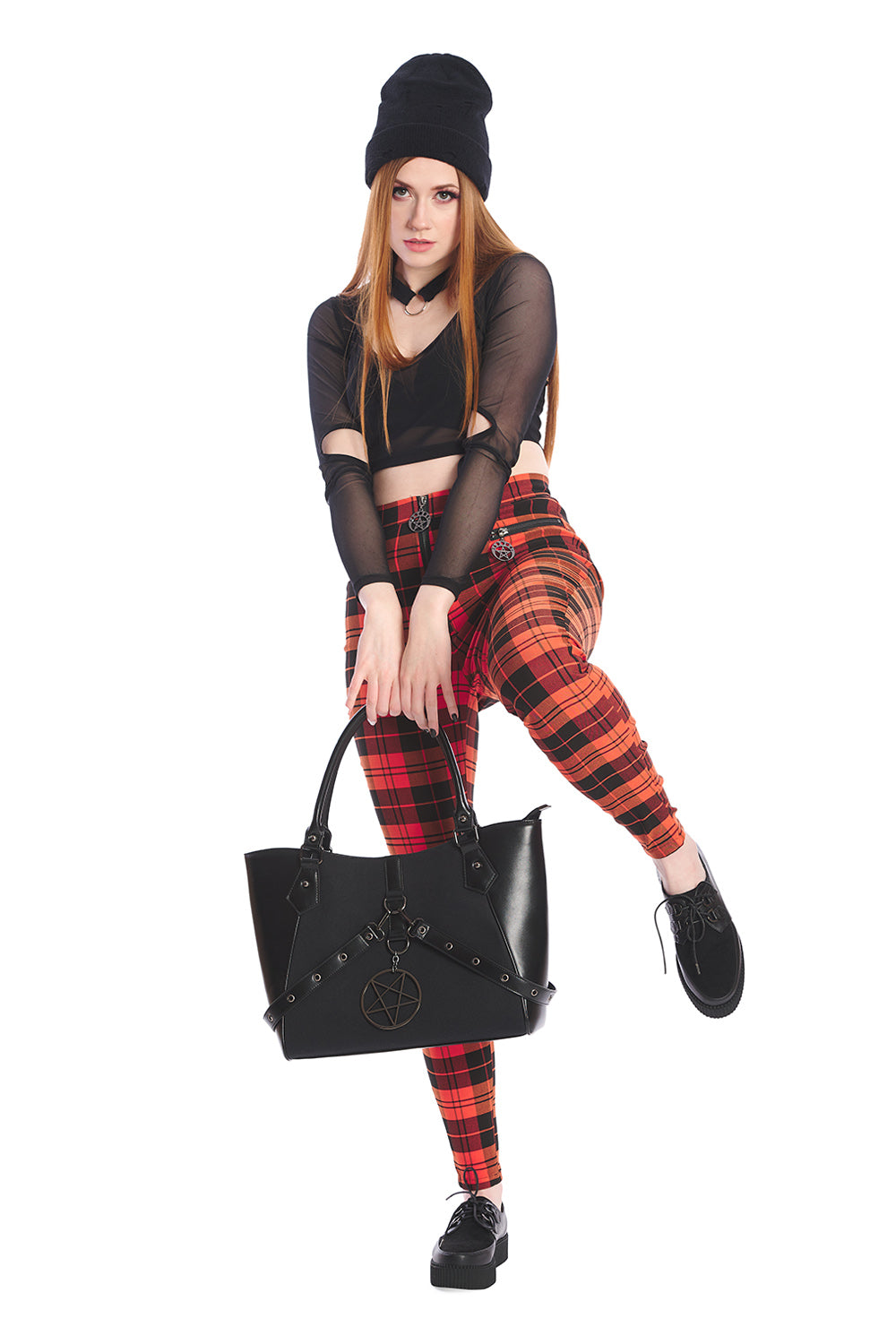 Model wears orange tartan high waisted trousers with a crop mesh top in black and beanie hat holding oversized handbag in black