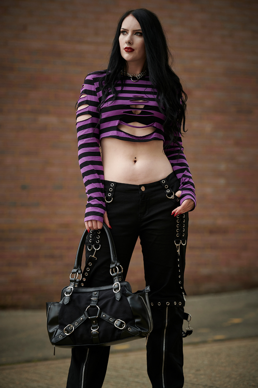 Lifestyle photo of Gothic model wearing a purple striped crop top with rip features and thumb hole sleeves holding a handbag with buckles 