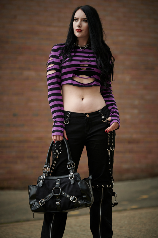 Lifestyle photo of Gothic model wearing a purple striped crop top with rip features and thumb hole sleeves holding a handbag with buckles